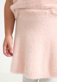Flauschiges pinkfarbenes Kleid mit weicher Textur und ausgestelltem Saum. Eine Hand hält den Rand, während graue Leggings darunter sichtbar sind.