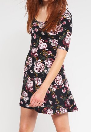 Femme portant une robe noire mi-longue à imprimé floral rose et blanc avec des manches mi-longues, debout devant un fond uni.