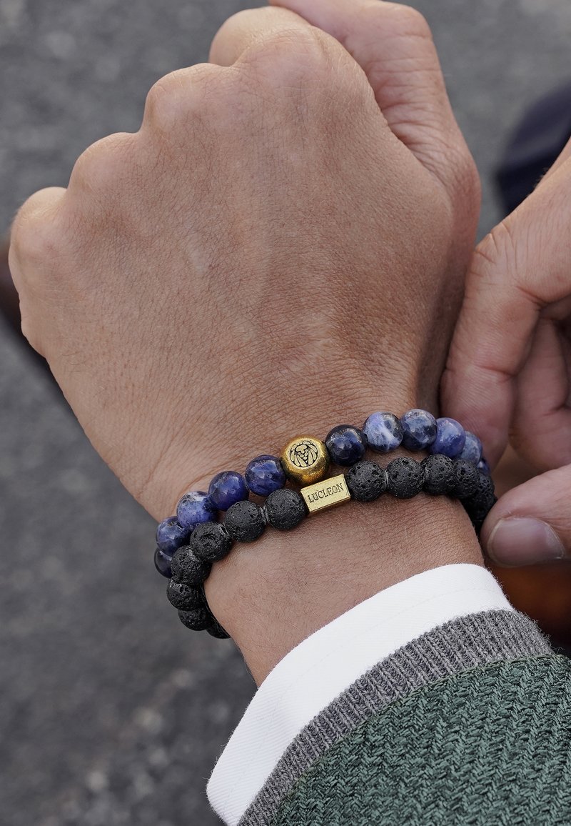 Armband aus Lavastein und blauer Sodalithperlen mit goldenen Akzenten, die ein Logo und ein rechteckiges Etikett zeigt. Tragen einer Person am Handgelenk, die einen grauen Pullover anhat.