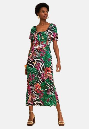 Žena v farebných midi šatách s kvetinovým a zebrovým vzorom, s nafúknutými rukávmi a béžovými sandálmi na vysokom podpätku.