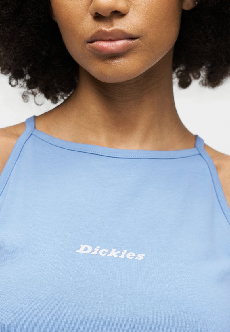 Giovane con capelli ricci che indossa una canottiera azzurro chiaro con la scritta "Dickies" in bianco sul petto.