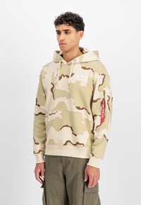 Sweatshirt à capuche kaki avec un motif camouflé crème et marron, poche frontale, et poche zippée avec un tirant rouge. Tissu en mélange de coton.