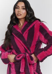Bata de baño de forro polar en rayas rosas y moradas oscuras, con cuello chal, cinturón anudado y bolsillos laterales. Tejido suave y texturizado.