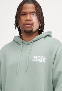 Groene hoodie van zacht materiaal, met een kangoeroe-pocket en koordjes. Witte "JACK & JONES" logo gedrukt op de borst.