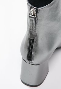 Gros plan d'une botte argentée à la cheville avec une fermeture éclair arrière et un talon bloc sur fond blanc.