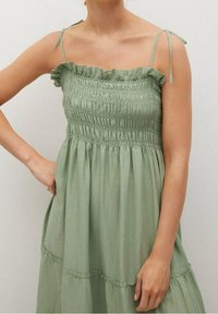 Femme portant une robe sans manches vert clair avec un corsage froncé et un col à volants noué aux épaules, sur un fond uni.
