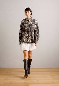 Camicia oversize con stampa leopardata e colletto abbinata a una gonna bianca e stivali neri di pelle alti fino al ginocchio. Sfondo in legno.
