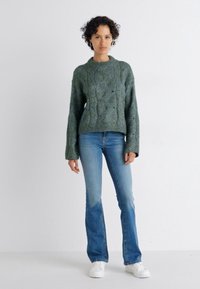 Maglione verde lavorato a maglia con un design a treccia testurizzata, abbinato a jeans svasati blu chiaro e sneakers bianche, su uno sfondo neutro.