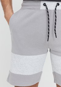 Shorts en mélange de coton gris avec des sections contrastantes de gris et de gris clair. Présentent une taille élastique avec des cordons de serrage noirs et des poches latérales.