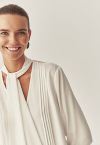 Biała bluzka z długim rękawem, o plisowanym wzorze i wiązaniu przy szyi. Wykonana z gładkiego materiału, z dekoltem w kształcie litery V i swobodnym krojem.
