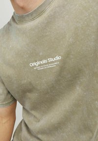 Oliven grøn t-shirt med et falmet look, som har en rund halsudskæring og korte ærmer. "Originals Studio" logo i hvid på forsiden.