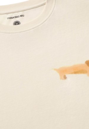 Camiseta color crema con un pequeño gráfico de un perro marrón claro cerca de la parte inferior derecha y etiqueta de la marca "THINKING MU" dentro del cuello.