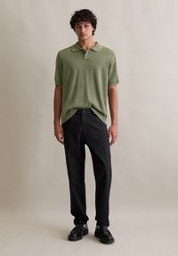 Groene gebreide polo met korte mouwen, afgeronde onderkant, gecombineerd met zwarte broeken en zwarte schoenen, staand tegen een neutrale achtergrond.