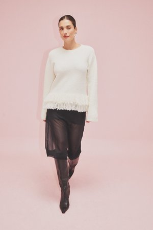 White textured sweater with fringe hem, paired with a sheer black skirt and knee-high black boots, set against a pink background.