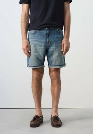 Mężczyzna w podwiniętych niebieskich szortach jeansowych, czarnym t-shircie i brązowych skórzanych mokasynach stoi na jednolitym tle.