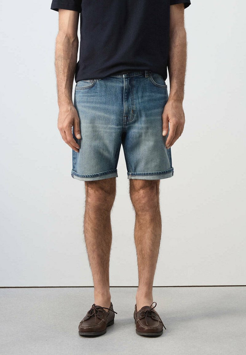 Homme portant un short en jean bleu retroussé, un t-shirt noir et des mocassins en cuir marron, debout devant un fond uni.