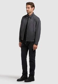 Homme debout portant une veste zippée gris foncé, un pantalon noir et des chaussures noires, sur un fond uni gris clair.