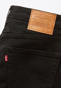 Vaqueros de mezclilla negros con un parche trasero de cuero que muestra "LEVI STRAUSS & CO." Etiqueta roja en la costura izquierda, costuras curvas en los bolsillos.
