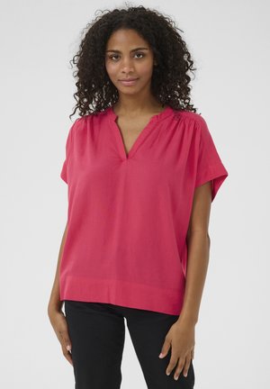 Blouse rose à manches courtes avec un décolleté en V, présentant une coupe ample et une texture subtile. Portée avec un pantalon noir ajusté, sur un fond uni.