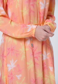 Pfirsichfarbenes Chiffonkleid mit floralem Muster in Rosa, Weiß und Gelb. Es hat elastische Bündchen und eine Kordelzug-Taille für eine verstellbare Passform.