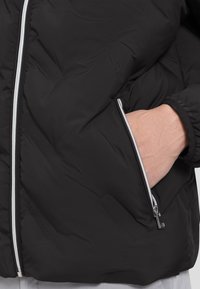 Eine Hand wird in die Reißverschlusstasche einer schwarzen Daunenjacke gesteckt, die einen kontrastierenden weißen Reißverschluss hat.