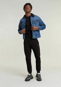 Giacca di jeans con collarino in pelliccia nera, felpa nera, pantaloni jogger neri e sneakers nere a fantasia. Il modello posa con sicurezza su uno sfondo neutro.