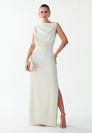 Donna che indossa un abito bianco senza maniche con uno spacco alto, tiene una pochette beige ed è accessoriata con orecchini, bracciale e tacchi dorati.