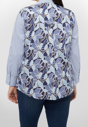 Femme portant une chemise à manches longues avec des manches rayées verticales bleu et blanc et un dos à motif floral bleu marine, associée à un jean foncé.