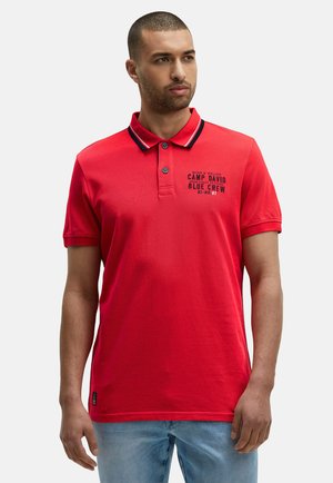 Mann in einem roten Poloshirt mit schwarz-weißer Borte am Kragen und bedrucktem Text auf der linken Brust, kombiniert mit hellblauen Jeans.