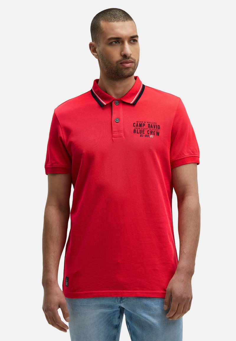 Uomo che indossa una polo rossa con rifiniture nere e bianche sul colletto e testo stampato sul lato sinistro del petto, abbinata a jeans azzurri chiari.