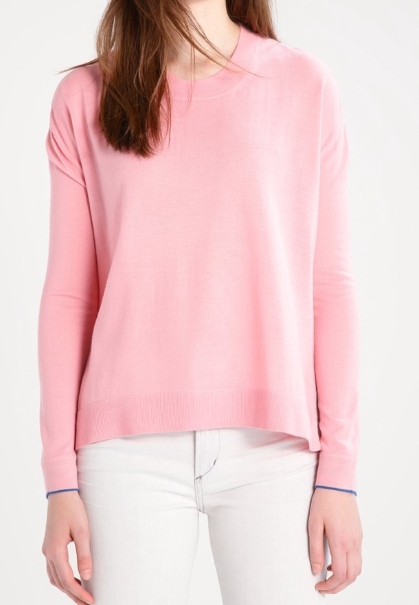 Personne portant un pull rose uni à manches longues avec poignets et ourlet côtelés, associé à un pantalon blanc sur fond blanc.