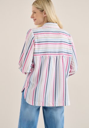 Vrouw met lang blond haar, gekleed in een losse, lange mouw, gestreepte blouse in roze, blauw en wit, gecombineerd met een blauwe spijkerbroek, zijwaarts gericht tegen een effen achtergrond.