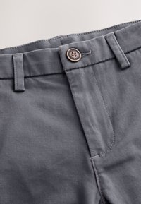 Primer plano de la cintura de unos pantalones grises con un botón etiquetado como "Authentic Next DUE" y presillas visibles sobre un fondo claro.