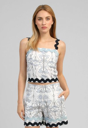 Femme aux cheveux blonds raides portant un haut blanc sans manches à motifs et un short assorti avec un bord ondulé noir et bleu, debout devant un fond gris.