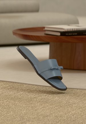 Sandale plate bleu à bout ouvert avec un petit nœud décoratif, flottant au-dessus d'un tapis beige texturé près d'une table basse en bois avec des livres empilés.