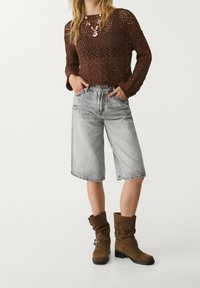 Suéter marrón de punto con mangas largas, pantalones cortos tipo bermuda de mezclilla gris, y botas marrones de caña alta con un detalle decorativo de hebilla.