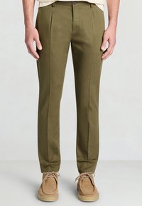 Olivgrüne Hose mit geradem Schnitt, zwei vorderen Falten und glatter Textur. Kombiniert mit hellbraunen Wildlederschuhen.