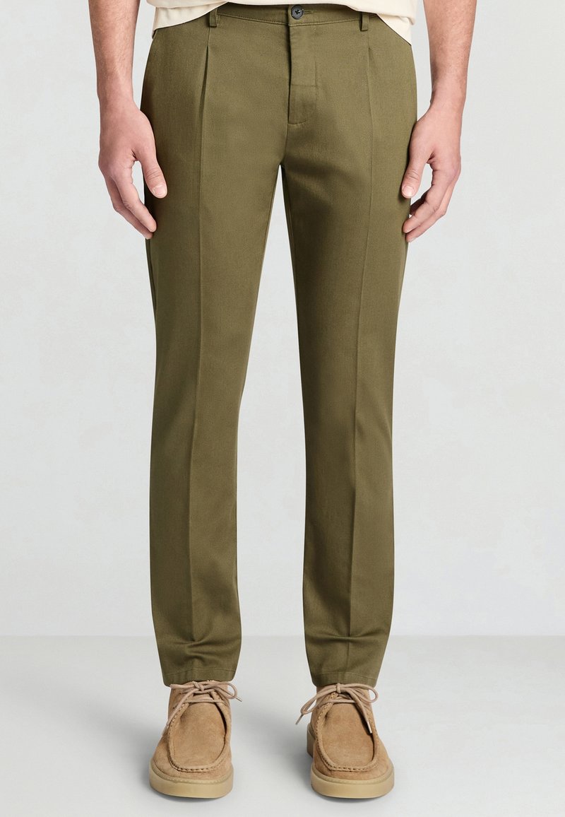 Olivgrüne Hose mit geradem Schnitt, zwei vorderen Falten und glatter Textur. Kombiniert mit hellbraunen Wildlederschuhen.