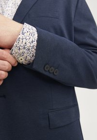 Granatowa marynarka z teksturowanej tkaniny, z trzema czarnymi guzikami na mankietach oraz jasną koszulą w kwiecisty wzór wystającą spod rękawa.