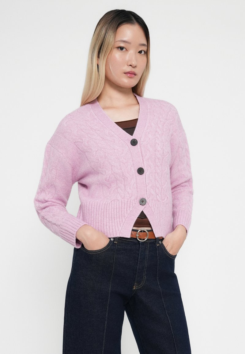 Cardigan en tricot rose avec un col en V, cinq boutons foncés, des manches et un ourlet côtelés, porté sur un haut marron et un jeans bleu foncé.