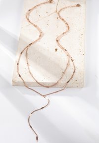 Naszyjnik warstwowy w kolorze różowego złota z różnymi długościami łańcuchów, z delikatnymi ogniwami i małymi akcentami, wyeksponowany na powierzchni teksturowanego kamienia.