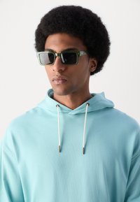 Occhiali da sole quadrati con montatura verde e lenti scure, indossati con una felpa leggera blu chiaro dotata di cordino e dettagli metallici.