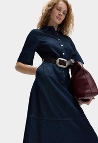 Mørkeblå denim skjortedress med korte ærmer, knaplukning foran og A-formet snit. Har et bredt sort bælte med en sølvspænde og en bordeaux taske.