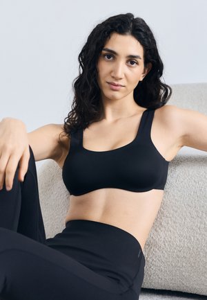 Donna con capelli ricci scuri che indossa un reggiseno sportivo nero e leggings, seduta rilassata su una poltrona beige testurizzata contro uno sfondo semplice.