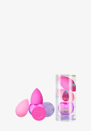 Trois éponges de maquillage roses de différentes formes et tailles, dont une avec une surface texturée, à côté d'un emballage cylindrique coloré.