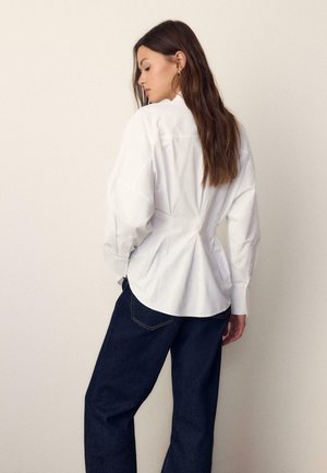 Ženska z dolgimi rjavimi lasmi, oblečena v belo oprijeto bluzo z dolgimi rokavi in temno modre kavbojke, stoji bočno ob preprostem svetlem ozadju.