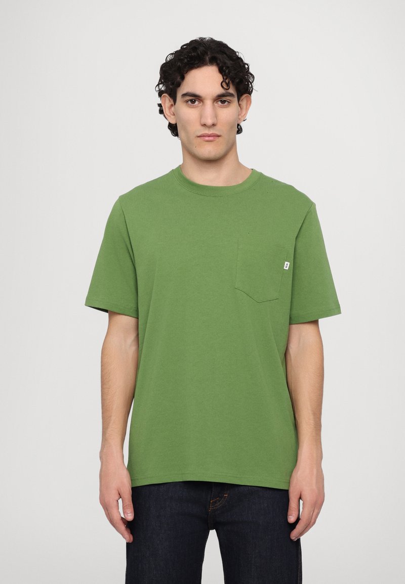 Green cotton T-shirt with short sleeves, crew neck, and a pocket on the left chest. Model wearing dark jeans stands against a plain background.