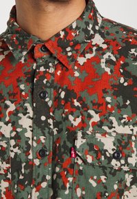 Corduroy overhemd in een groen, rood en wit camouflagepatroon; heeft een knoopsluiting bij de kraag, twee opgestikte zakken en zwarte knopen.