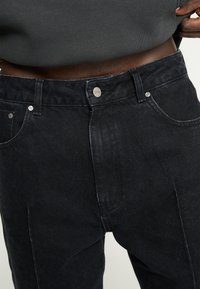 Nærbillede af en person iført højtaljede sorte jeans med metalknapper og nitter, der trækker en mørk skjorte op for at afsløre deres talje.