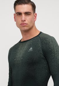 Majica dugih rukava za sport u tamnozelenoj boji, s teksturiranim uzorkom, prilagođenog kroja, okruglog izreza, s malim bijelim logotipom na prsima.